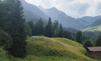 A picturesque landscape with green hills and surrounded by tall mountains. In the foreground stands a small cabin, surrounded by trees. | © Nicole Lughammer