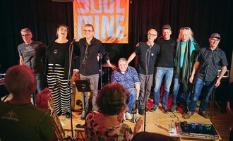 A band stands on stage and smiles at the camera. In the background, there is a bright banner with the inscription "SOUL". | © Soulmine