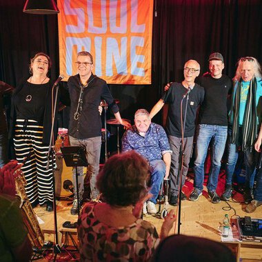 A band stands on stage and smiles at the camera. In the background, there is a bright banner with the inscription "SOUL". | © Soulmine