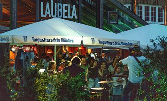 Eine lebendige Atmosphäre unter einem Zelt mit der Aufschrift "Augustiner-Bräu München". Leute genießen Musik und Gemeinschaft in einem belebten Außenbereich. | © Auenhütte | Basti Heckl