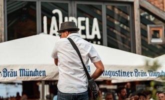 Eine lebhafte Straßenszene mit einer Menschenmenge, die einem Performenden zuschaut. Im Hintergrund ist das Restaurant „La Capela“ zu sehen. | © Auenhütte | Basti Heckl