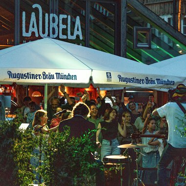 Eine lebendige Atmosphäre unter einem Zelt mit der Aufschrift "Augustiner-Bräu München". Leute genießen Musik und Gemeinschaft in einem belebten Außenbereich. | © Auenhütte | Basti Heckl