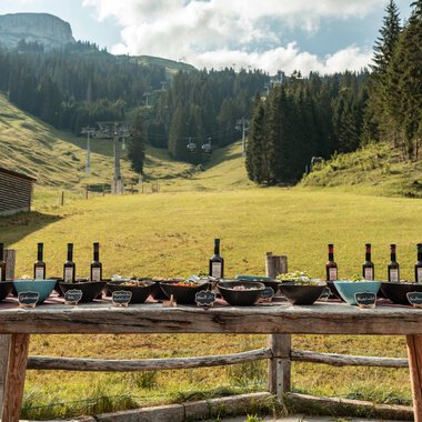 Ein schöner Tisch im Freien, gedeckt mit Weinflaschen und verschiedenen Speisen. Im Hintergrund sind sanfte Hügel und Bäume zu sehen. | © Auenhütte | Basti Heckl