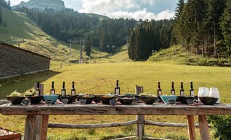 Ein schöner Tisch im Freien, gedeckt mit Weinflaschen und verschiedenen Speisen. Im Hintergrund sind sanfte Hügel und Bäume zu sehen. | © Auenhütte | Basti Heckl