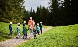 Eine Gruppe von Kindern mit Helmen wandert einen Weg entlang, begleitet von einer Erwachsene. Die Umgebung besteht aus grünen Wiesen und hohen Bäumen. | © Kleinwalsertal Tourismus | Oliver Farys