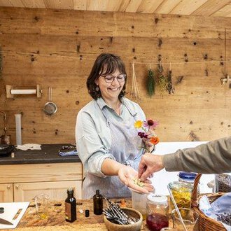 Eine lächelnde Frau in einer Holzküche reicht einer Person Blumen und kleine Flaschen. Im Hintergrund sind Kräuter und verschiedene Zutaten zu sehen. | © Biohotel Walserstuba | Patrick Säly