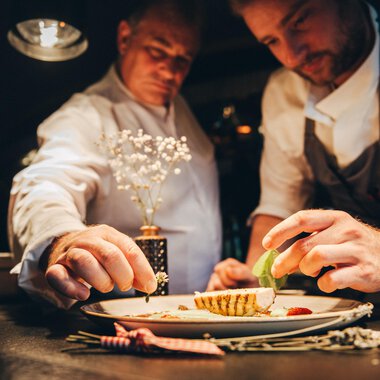 Zwei Köche, die an einem Teller arbeiten, während sie sorgfältig Zutaten hinzufügen. Die Atmosphäre ist warm und einladend, mit einem Fokus auf das kunstvolle Anrichten des Gerichts. | © Hotel Birkenhöhe Bantel GmbH & Co KG