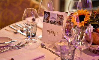 Ein elegant gedeckter Tisch mit Gläsern und Besteck. In der Mitte steht eine Menükarte und ein Blumengesteck mit Sonnenblumen. | © Hotel Birkenhöhe | Annette Sandner