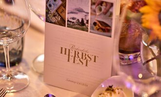 Ein elegant gedeckter Tisch mit Gläsern und einer Speisekarte für das Herbstfest. Im Hintergrund sind Blumen und eine kleine Schale mit einer delicaten Speise zu sehen. | © Hotel Birkenhöhe | Annette Sandner