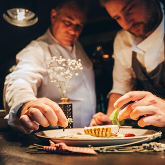 © Hotel Birkenhöhe Bantel GmbH & Co KG Two chefs working on a plate while carefully adding ingredients. The atmosphere is warm and inviting, with a focus on the artistic presentation of the dish. | © Hotel Birkenhöhe Bantel GmbH & Co KG