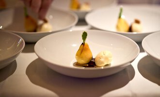 Ein schön angerichteter Teller mit einer aufrecht stehenden Birne, serviert mit einer Kugel Eis und einer grünen Dekoration. Der Hintergrund zeigt mehrere ähnliche Teller. | © Hotel Birkenhöhe | Annette Sandner