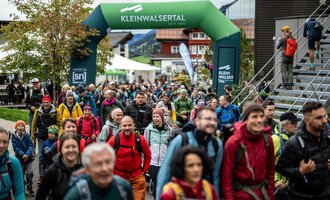 Two hikers walk along a path in the mountains. The sun is shining and the landscape is green and golden. | © Kleinwalsertal Tourismus eGen - Fotograf: Oliver Farys