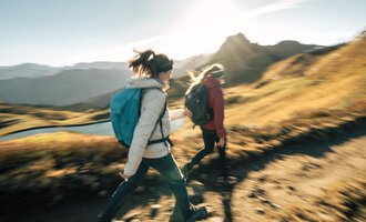 Two hikers walk along a path in the mountains. The sun is shining and the landscape is green and golden. | © Kleinwalsertal Tourismus eGen - Fotograf: Hannah Bichay
