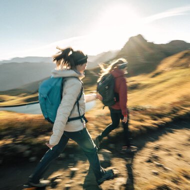 Two hikers walk along a path in the mountains. The sun is shining and the landscape is green and golden. | © Kleinwalsertal Tourismus eGen - Fotograf: Hannah Bichay