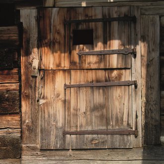 © R. Koeberle Eine alte Holzlatttür mit Eisenbeschlägen. Die Tür hat ein kleines Fenster und ist von einer rustikalen Wand umgeben. | © R. Koeberle