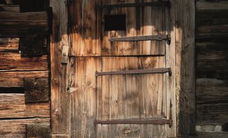 Eine alte Holzlatttür mit Eisenbeschlägen. Die Tür hat ein kleines Fenster und ist von einer rustikalen Wand umgeben. | © R. Koeberle