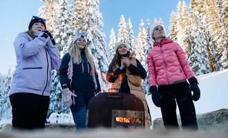 Eine Gruppe von Frauen steht um einen kleinen Heizofen in einer verschneiten Winterlandschaft. Im Hintergrund sind schneebedeckte Bäume und ein klarer, blauer Himmel zu sehen. | © Kleinwalsertal Tourismus | Oliver Farys