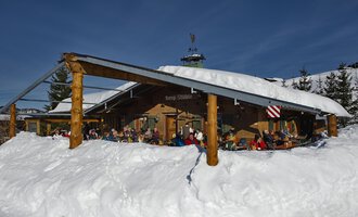 Eine gemütliche Berghütte im Schnee mit vielen Menschen auf der Terrasse. Die Umgebung ist winterlich und die Sonne scheint am blauen Himmel. | © Bergstüble | Michaela Beck Dornach