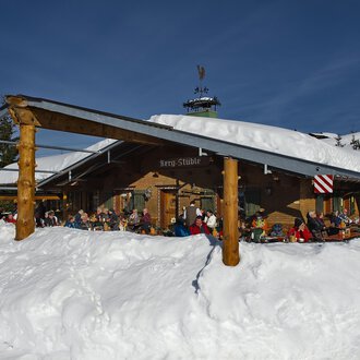 Eine gemütliche Berghütte im Schnee mit vielen Menschen auf der Terrasse. Die Umgebung ist winterlich und die Sonne scheint am blauen Himmel. | © Bergstüble | Michaela Beck Dornach
