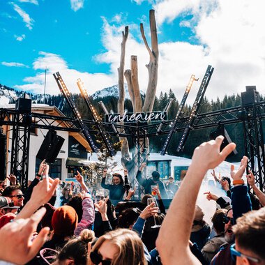 A lively festival atmosphere with a large crowd dancing to music. In the background, a stage and snow-covered mountains are visible.