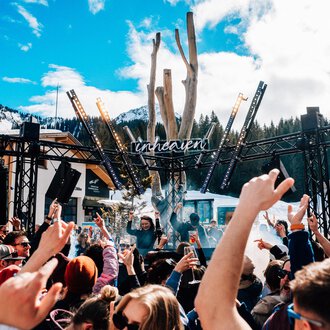 A lively festival atmosphere with a large crowd dancing to music. In the background, a stage and snow-covered mountains are visible.