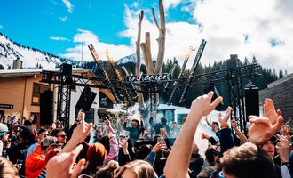 A lively festival atmosphere with a large crowd dancing to music. In the background, a stage and snow-covered mountains are visible.