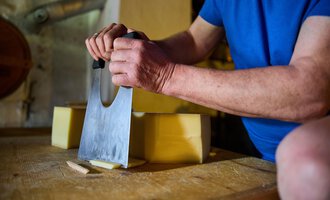 Eine Person schneidet Käse mit einem großen Messer auf einem Holztisch. Die Szene zeigt handwerkliches Geschick und Frische der Zutaten. | © Kleinwalsertal Tourismus eGen - Fotograf: Oliver Farys