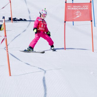 Ein kleiner Skifahrer in pinker Skiausrüstung fährt durch ein Tor auf einer verschneiten Piste. Im Hintergrund sind weitere Skifahrer und Farbmarkierungen zu sehen.