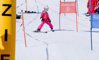 A small skier in pink ski gear is skiing through a gate on a snowy slope. In the background, other skiers and color markings can be seen.