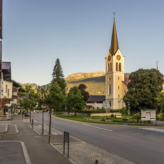 Eine ruhige Straße mit einem kleinen Dorf und einer Kirche im Vordergrund. Im Hintergrund sind grüne Bäume und Berge zu sehen. | © Kleinwalsertal Tourismus | Steffen Berschin