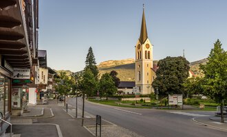 Eine ruhige Straße mit einem kleinen Dorf und einer Kirche im Vordergrund. Im Hintergrund sind grüne Bäume und Berge zu sehen. | © Kleinwalsertal Tourismus | Steffen Berschin