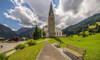 A picturesque church with a tall tower in a green landscape. In the background, mountains and a cloudy sky are visible. | © Kleinwalsertal Tourismus | Steffen Berschin