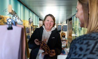 Eine lächelnde Frau zeigt ein Accessoire einer anderen Person. Im Hintergrund sind weitere Produkte in einem hellen, modernen Verkaufsraum zu sehen. | © Kleinwalsertal Tourismus | Oliver Farys