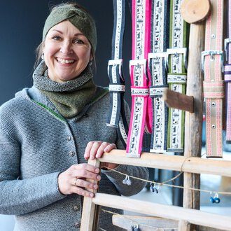 Eine freundlich lächelnde Frau steht vor einem Ständer mit bunten Bändern. Sie trägt einen grauen Pullover und einen Schal. | © Kleinwalsertal Tourismus | Oliver Farys