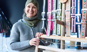 Eine freundlich lächelnde Frau steht vor einem Ständer mit bunten Bändern. Sie trägt einen grauen Pullover und einen Schal.