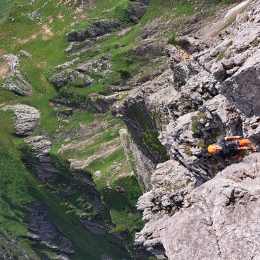An impressive mountain landscape with steep cliffs and green meadows. A climber is on a rock face. | © stoamandl.at | Olli Sinz