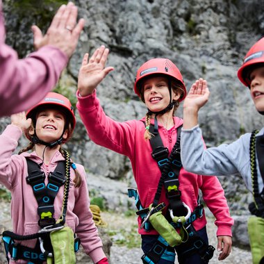 Drei Kinder in Kletterausrüstung begrüßen einen Erwachsenen. Sie tragen Helme und lächeln vor einer felsigen Kulisse. | © Kleinwalsertal Tourismus | Oliver Farys