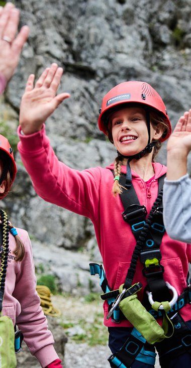 Drei Kinder in Kletterausrüstung begrüßen einen Erwachsenen. Sie tragen Helme und lächeln vor einer felsigen Kulisse. | © Kleinwalsertal Tourismus | Oliver Farys