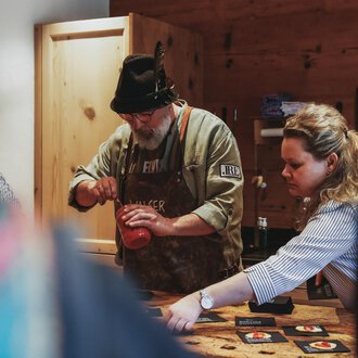A man and a woman are preparing something in a cozy kitchen. The man is wearing a hat and an apron, while the woman is working intently. | © Angela Lamprecht | Vorarlberg Tourismus