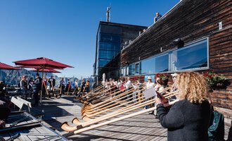 Eine Gruppe von Menschen spielt auf Alphörnern auf einer Terrasse. Im Hintergrund sind sonnige Berge und eine klare blaue Himmel zu sehen. | © Kleinwalsertal Tourismus | Berthold Schick