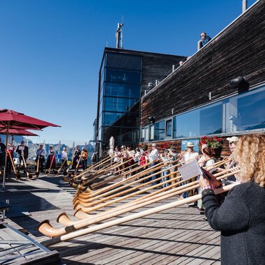 Eine Gruppe von Menschen spielt auf Alphörnern auf einer Terrasse. Im Hintergrund sind sonnige Berge und eine klare blaue Himmel zu sehen. | © Kleinwalsertal Tourismus | Berthold Schick