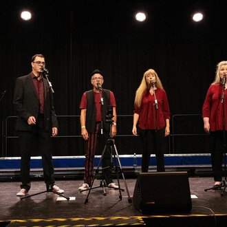 © Nice Try Eine Gruppe von sechs Sängern steht auf einer Bühne und singt in Mikrofone. Alle tragen rot und schwarz und die Bühne ist schwach beleuchtet. | © Nice Try