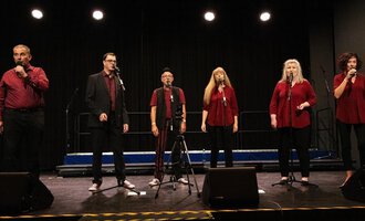 Eine Gruppe von sechs Sängern steht auf einer Bühne und singt in Mikrofone. Alle tragen rot und schwarz und die Bühne ist schwach beleuchtet. | © Nice Try