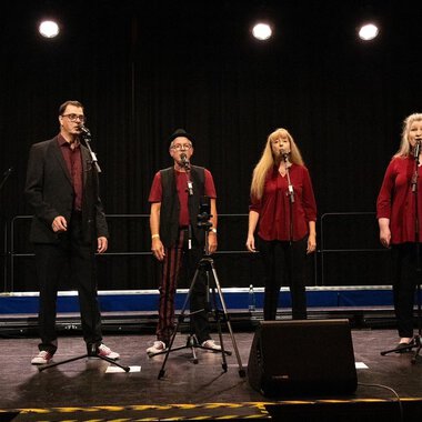 Eine Gruppe von sechs Sängern steht auf einer Bühne und singt in Mikrofone. Alle tragen rot und schwarz und die Bühne ist schwach beleuchtet. | © Nice Try