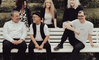 A group of six people is sitting on a bench outdoors. They are laughing and seem to be having a good time together. | © Nice Try