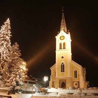 Eine verschneite Landschaft mit einer beleuchteten Kirche und einem geschmückten Weihnachtsbaum. Der Nachthimmel verstärkt die festliche Atmosphäre. | © Kleinwalsertal Tourismus | Frank Drechsel