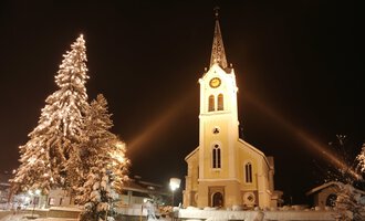 Eine verschneite Landschaft mit einer beleuchteten Kirche und einem geschmückten Weihnachtsbaum. Der Nachthimmel verstärkt die festliche Atmosphäre. | © Kleinwalsertal Tourismus | Frank Drechsel