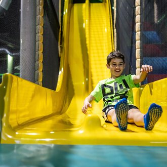© Wonnemar Sonthofen | interSPA Gruppe A smiling child happily slides down a yellow slide. In the background, playground equipment can be seen. | © Wonnemar Sonthofen | interSPA Gruppe