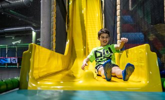 A smiling child happily slides down a yellow slide. In the background, playground equipment can be seen. | © Wonnemar Sonthofen | interSPA Gruppe