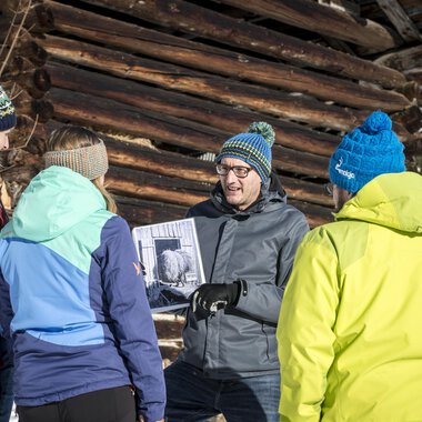 A group of five people stands in the snow in front of a wooden barn. They are collectively looking at a photo that one of the participants is holding in their hand. | © Kleinwalsertal Tourismus | Dominik Berchtold